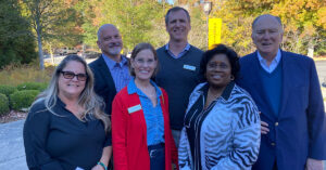 Board members on campus in front of trees and gold university banner